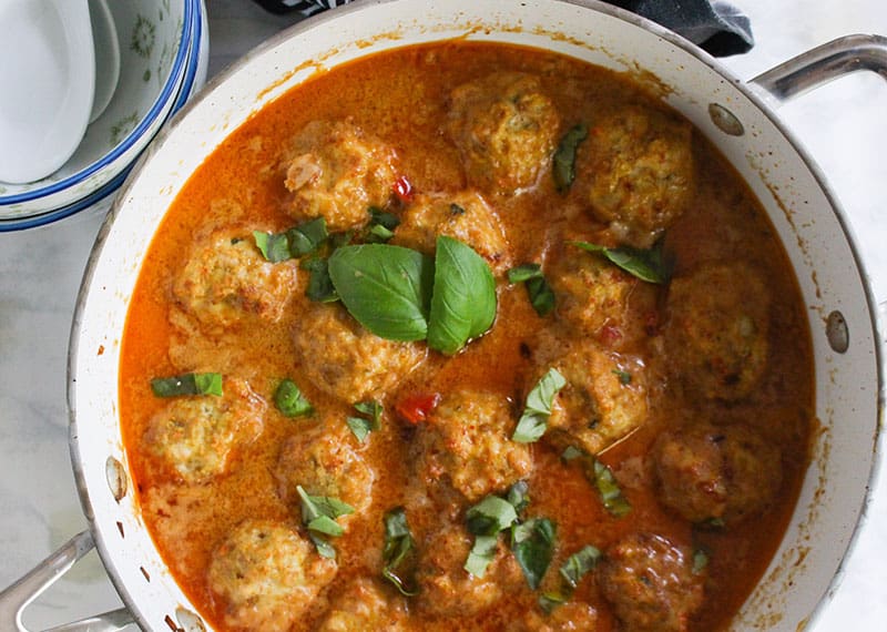 A large pot is full of chicken meatballs in a coconut curry sauce. There are green garnishes sprinkled across the top.