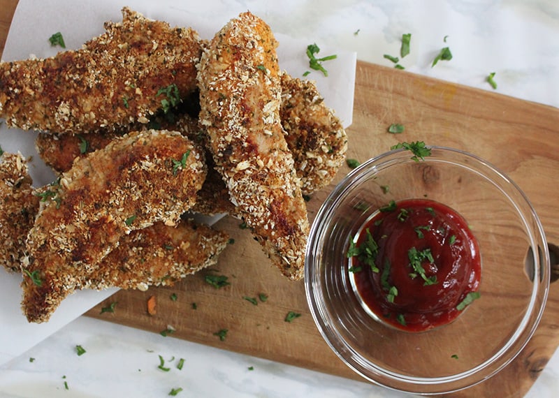 Oat Crusted Chicken Tenders lay atop a wooding cutting board. A glass dish with a dipping sauce is to the right of the tenders