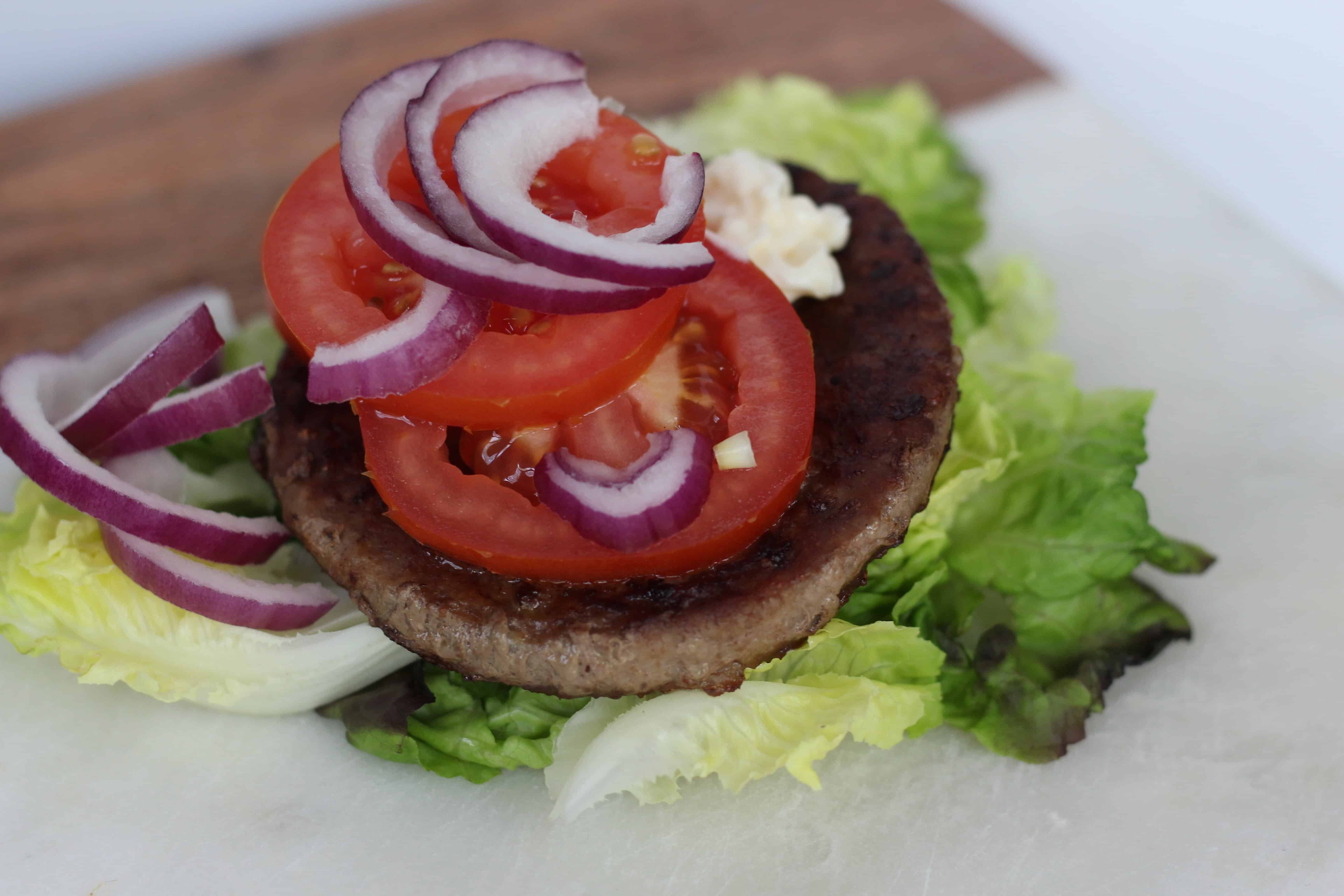 A burger patty topped with a red tomato, feta cheese and red onion lies atop romaine lettuce