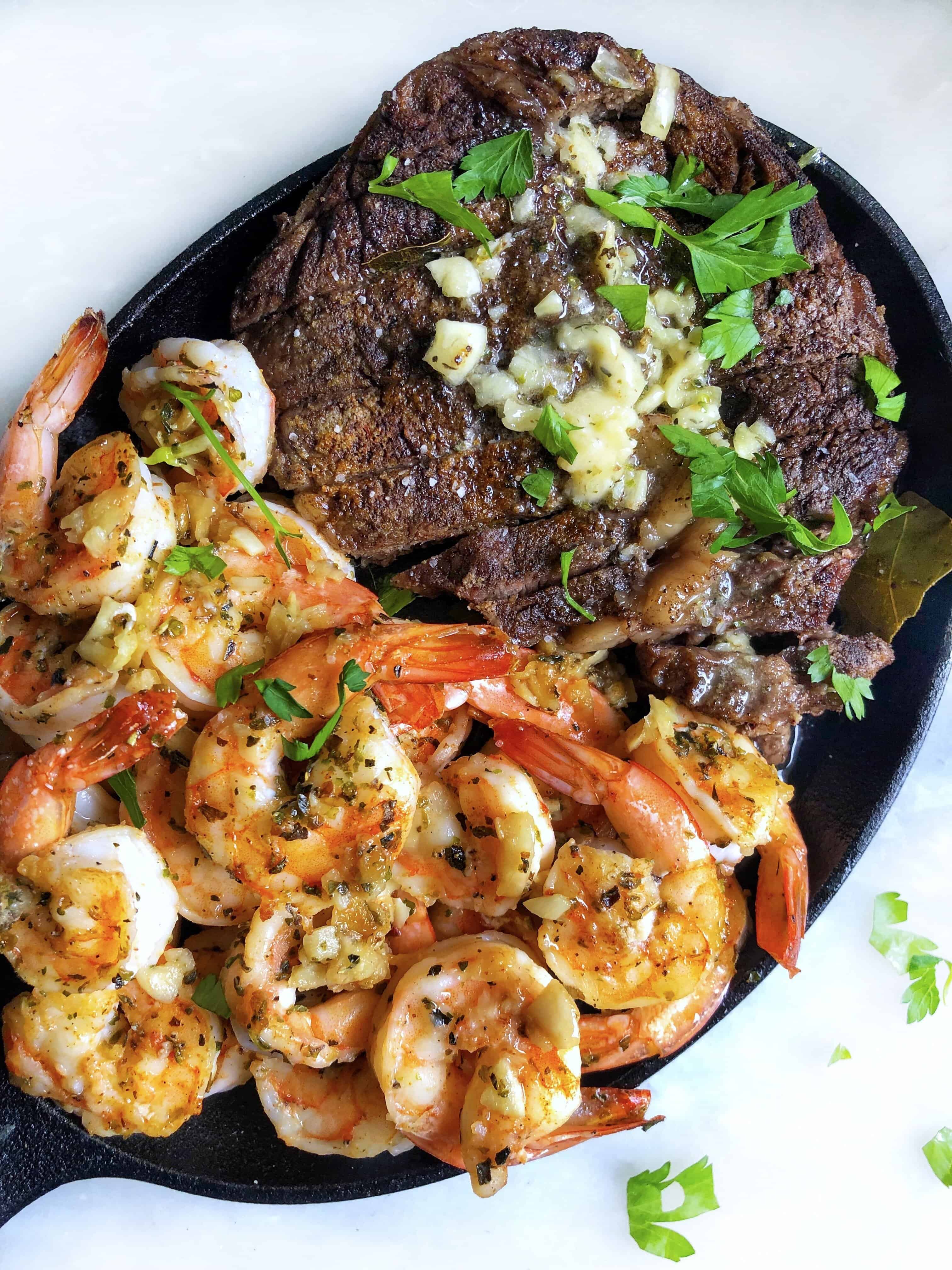 A black pan lays on a counter top. One half of the pan contains shrimp, the other half contains steak.
